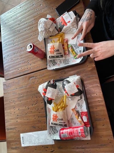 The scene depicts two fast-food meals placed on a rustic, textured wooden table within the CHB Burger restaurant in Plaisir, France, as identified by "CHB" branding on packaging and receipts, and "Kiosk 325" on order labels.

Two black plastic trays each contain elements of a meal. The top tray holds one burger wrapped in branded paper, a white paper bag of french fries with "#CHB" logo, and two sauce packets (one red, one green/yellow). Adjacent to this tray on the table is an open red aluminum can, partially identified as a Coca-Cola product, and a black smartphone lying face down. The bottom tray contains two burgers wrapped in branded paper, a white paper bag of french fries with "#CHB" logo, and two sauce packets (one red, one brown/gold). An open red aluminum can of Coca-Cola Zero Sugar is positioned on this tray. A long white receipt with "#CHB" branding rests on the table to the left of the bottom tray.

The hands of one individual are visible on the right side of the image. The left hand displays extensive, dark, intricate tattoos covering the back of the hand and fingers. The right hand wears a silver ring on the ring finger. Both hands are posed in a "peace sign" gesture. The individual wears dark clothing, with a sleeve visible. The meals appear prepared for consumption.