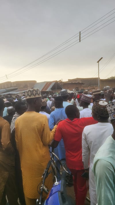 This image depicts a large gathering of men outdoors in Jogana, Nigeria. The scene is illuminated by a soft, warm light, suggesting either early morning or late afternoon under a clear sky. Most of the men are dressed in traditional Muslim attire, including long robes (kaftans or boubous) and distinctively embroidered prayer caps (kufis or tagiyas). Some individuals are seen in casual wear, such as a red shirt and a blue shirt, adding variety to the crowd.

The men appear to be seated or standing attentively, with their gazes directed towards a common point outside the frame, indicating participation in a communal event. This posture and traditional dress strongly suggest a religious gathering, such as congregational prayers or a sermon.

The setting features simple, low-rise buildings with mud-brick walls and corrugated metal roofs in the background, typical of a rural or semi-urban Nigerian community. A utility pole with multiple overhead power lines cuts across the sky in the upper left. In the foreground, parts of a blue motorbike/scooter and a bicycle handlebar are visible. The atmosphere appears peaceful and orderly. No specific emotions are clearly visible on individual faces. There is no visible text within the image.
