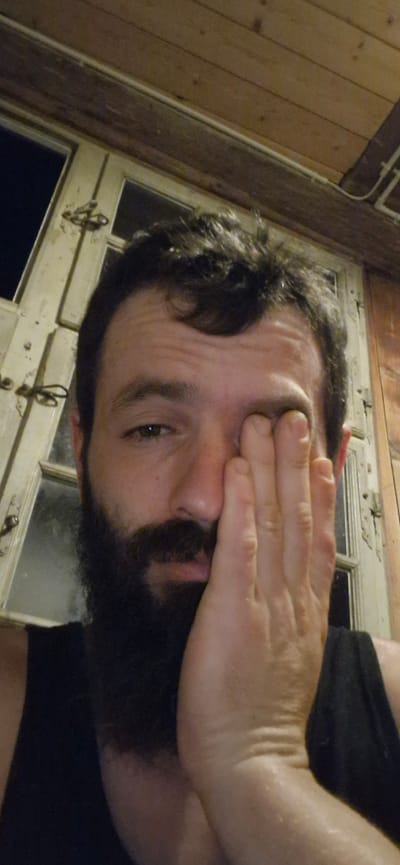 A medium close-up shot is captured from a low angle, focusing on the face of a man with a beard and dark, curly hair. He is holding his right hand over his right eye, with his fingers splayed. His left eye is visible and appears to be looking forward. The background consists of a wooden ceiling and a window with cream-colored panes and dark hardware. The man is wearing a black tank top. The overall scene suggests an indoor setting, possibly a room or a covered outdoor space, with elements of rustic architecture. The image was taken in Wattenwil, Switzerland.