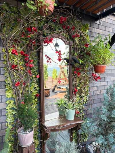 In Nizhnyaya Shilovka, Russia, a decorative outdoor scene features a wooden-framed mirror surrounded by artificial vines and red artificial flowers, attached to a dark grey brick wall. A black crow figurine is perched on the left side of the mirror's vine decoration. Two potted plants, one with green leaves in a light blue pot and another with striped leaves in a sage green pot, sit on a wooden shelf below the mirror. To the right of the mirror, a larger potted plant with long green leaves hangs from a terracotta pot. In the foreground, a small evergreen plant in a pink textured pot is positioned on a dark wooden stand. A cactus plant is visible to the left, and a blue-green conifer tree stands to the right, with a small pot filled with stones in front of it. The mirror reflects a wooden deck, a swing set, and a mountainous landscape under a cloudy sky. The scene is covered by a wooden roof structure with visible beams.
