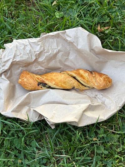 An eye-level shot depicts a chocolate twist pastry on crumpled brown paper, resting on grass. The scene suggests an outdoor setting in a natural area, likely a park, based on the grass and clover. There are no people or explicit indications of a location within the city of High Wycombe, United Kingdom, other than potential environmental factors (such as the types of plants) that are consistent with a British climate. The pastry is the focal point, surrounded by the crumpled paper and the grass.
