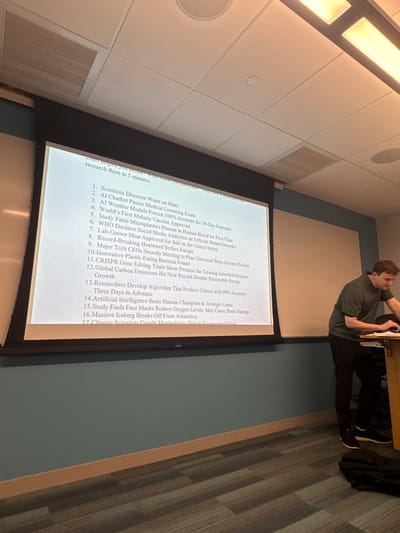 The image depicts the interior of a modern classroom or lecture hall, located in Waltham, United States. A large white projector screen occupies the left half of the frame, displaying a numbered list of seventeen diverse topics, including "Scientists Discover Water on Mars," "AI Chatbot Passes Medical Licensing Exam," and "World's First Malaria Vaccine Approved." Text above the list indicates an instruction to "research them in 5 minutes." To the right of the screen, a young adult male with short dark hair, wearing a dark green polo shirt and dark trousers, stands at a light wood-toned lectern. He is leaning slightly forward, with his hands positioned on the lectern, suggesting interaction with a device such as a laptop. The room features a white drop ceiling with rectangular fluorescent light fixtures and air vents. The lower portion of the walls is painted a teal-blue color, transitioning to a lighter shade higher up, while the floor is covered with a patterned grey carpet.