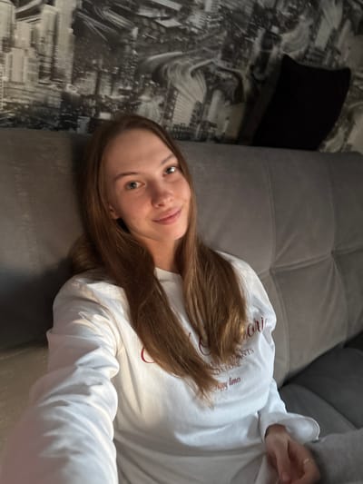 A young woman with long, light brown hair is captured in a medium shot, seated on a gray couch. She is wearing a long-sleeved white sweatshirt with red lettering. Her gaze is directed towards the camera, and she has a slight smile. Behind her, a wall is decorated with a black and white city skyline mural. A dark pillow is visible on the couch to her right. The lighting suggests an indoor environment, possibly in the evening. The image was taken in Novaya Ivanovka, Russia.