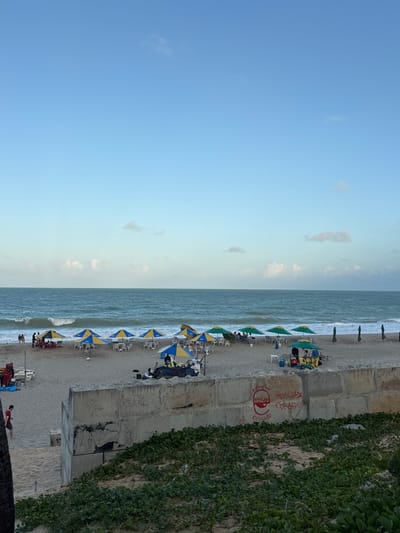 An outdoor wide shot captures a populated beach scene in Natal, Brazil, under a clear light blue sky with sparse white cumulus clouds. The deep blue ocean presents moderate white-capped waves breaking towards the shore, with a discernible horizon line. The sandy beach is occupied by approximately 15-20 individuals. Many are seated under 10-12 blue and yellow, and 5-7 green beach umbrellas, each accompanied by white plastic chairs. Several vendor setups are visible on the sand, including one with a blue and yellow umbrella and another with a green umbrella, featuring coolers and tables. On the far left, two white small boat-like structures and blue tarpaulins are present. A low, weathered concrete wall, stained and marked with graffiti, runs across the mid-foreground, separating the beach from an elevated sandy dune covered in dense green ground vegetation. Red graffiti on the concrete wall, from left to right, includes the faint word "REAL," a stylized red logo, "Sempre Humildade Coração," and "Obrigado." A dark tree trunk is partially visible on the far left foreground, obscuring the lower left corner. One individual in a red shirt and dark shorts is observed walking on the sand in the lower-left quadrant.