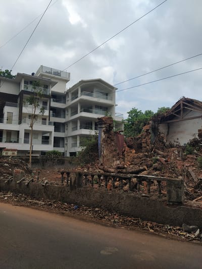 The image shows a modern white building next to a collapsed or partially demolished brick structure in what appears to be an urban setting. The building has multiple stories, with balconies and large windows. Adjacent to it, a pile of rubble, broken bricks, and debris suggests recent demolition or collapse of an older brick structure. The scene is situated next to a paved road, and overhead power lines are visible in the sky. The sky is overcast with clouds. The general location may be Marna, India as indicated in the prompt. There are no people visible in the shot.