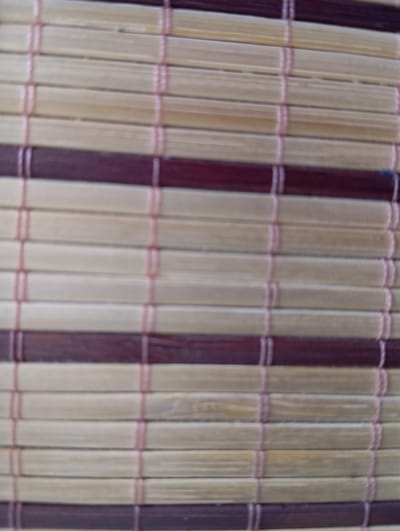 The image shows a close-up, slightly blurred view of a woven bamboo mat. The mat is made of horizontal strips of light-colored bamboo alternating with darker, reddish-brown bamboo strips. These strips are held together by thin, pinkish threads that run vertically. The texture is rough and organic, with visible grain patterns on the bamboo. The lighting appears to be natural and diffuse, casting no harsh shadows. The overall impression is one of a rustic, textured surface. There is no identifiable location in the city of Hlynytsi, Ukraine, or any people, actions, or interactions present in the image.