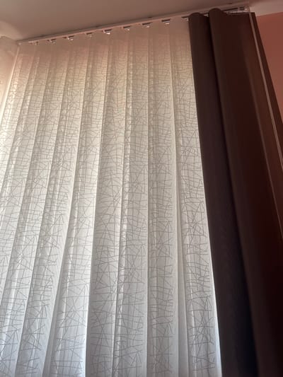 The image shows a close-up of a dual-layer window blind. The top layer is a dark brown, opaque fabric that appears to be a valance or a rolled-up section of the blind. Below this is a patterned translucent fabric, consisting of horizontal stripes. These stripes alternate between a sheer material and a material with a delicate, web-like pattern of thin, interconnected lines. The blind is installed in what appears to be an indoor setting, likely a room with natural light filtering through the translucent sections. The lighting suggests it could be daytime. No people, specific activities, or discernible text are visible. The setting is likely a residential or commercial interior in Branipole, Bulgaria.