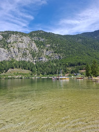 The image depicts a scenic view of Lake Hallstatt in Obertraun, Austria, on a sunny day with a partly cloudy sky. The foreground features the clear, shallow water of the lake, revealing the pebbled lakebed. In the middle ground, several sailboats are docked at a wooden pier, with one small red boat further out on the water. Along the shore, a cluster of buildings, likely homes or businesses, are nestled amongst trees. The background is dominated by a majestic mountain range covered in dense evergreen forest, with rocky outcrops visible on the steeper slopes. The overall impression is one of a tranquil, picturesque alpine landscape.