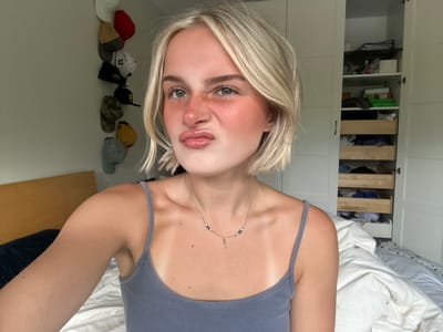 The media file is a portrait-oriented selfie taken indoors. The main subject is a young person with blonde hair, who is facing the camera with their head tilted upside down. They are wearing a grey tank top and a delicate necklace. Their facial expression is playful, with bright eyes and slightly parted lips. The background shows an open closet with shelves filled with clothing and various items, as well as a wall with a row of baseball caps. The lighting suggests it is daytime. The presence of a closet and the caps indicates a casual, domestic setting. There is no visible text in the image.