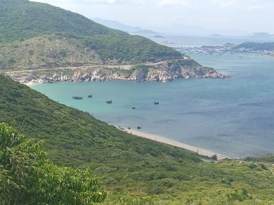 This image captures a panoramic view of a coastal landscape in Vinh Hai, Vietnam. The foreground is dominated by a lush, green hillside covered in dense vegetation, suggesting a tropical environment. A winding road snakes along the side of a rocky, tree-covered cliff face overlooking a tranquil turquoise bay. In the bay, several small boats are anchored, their forms silhouetted against the calm water.
Further out, the bay opens up to a wider expanse of ocean, where a distant coastline is visible. This coastline is dotted with a cluster of buildings, likely a town or village, and a much larger concentration of boats, indicating a busy harbor. The horizon is hazy, with soft blue mountains fading into the pale sky, suggesting a bright, clear day. The overall scene conveys a sense of natural beauty and coastal life.