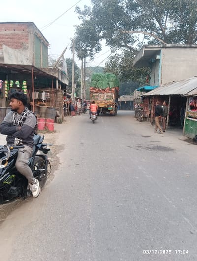 This image depicts a late afternoon street scene in Maliat, Bangladesh, dated 03/12/2025 at 17:04. The primary subject in the foreground, left of center, is a young male with dark hair and a beard, wearing a grey and black patterned long-sleeve shirt, grey pants, and white sneakers. He is seated on a black sport-style motorcycle, identified by a "310" decal, with his right foot resting on the ground, facing left away from the viewer. The motorcycle is positioned on the unpaved shoulder of an asphalt road that extends into the distance.
In the midground, a yellow truck heavily loaded with goods, including orange sacks and items covered by a green tarp, proceeds away from the viewer in the center of the road. Behind it, a person wearing an orange shirt is riding a red motorcycle. On the left side of the road, a series of small, makeshift market stalls or shops with corrugated metal roofing and brick structures are visible. Red gas cylinders are stacked near the foreground motorcycle on the left. Clothing items hang from displays in one stall. Multiple indistinct individuals are visible near these stalls further down the street. On the right side of the road, another line of similar small stalls with corrugated metal roofs is present. Two men are standing near one of these stalls, observing or interacting.
The background shows numerous trees with sparse foliage, suggesting a dry or winter season, alongside more buildings and utility poles with overhead wires. The sky is bright but hazy or overcast, indicating late afternoon conditions. The road surface appears worn.