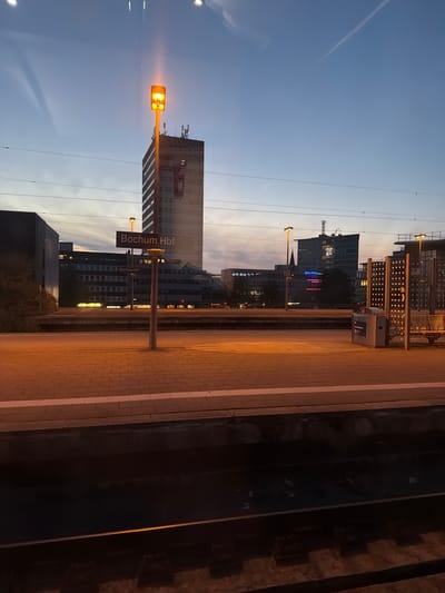 This media file shows a view of the Bochum Hauptbahnhof (main train station) in Bochum, Germany. The image is oriented vertically, with the sky at the top and the train tracks/platform at the bottom.

The subjects are primarily architectural and environmental. We see a tall, modern building with many windows, reflecting the sky and surrounding lights. In the foreground, a train station platform with a tiled surface and white lines is visible. A street lamp with a warm, orange glow is prominently featured, illuminating the platform. Further in the background, more buildings are visible, some with lit windows, suggesting it's dusk or evening. There are also overhead power lines for the trains.

The scene depicts a train station setting, likely during the transition from day to night. The lighting suggests either late afternoon or early evening. The sky is a pale blue, with some lighter areas that could indicate clouds or the setting sun.

Notable details include the "Bochum Hbf" sign on a pole, clearly indicating the location. Another, smaller sign with the same text is visible on a building in the background. The overall impression is one of urban infrastructure and the quiet atmosphere of a train station at this time of day. There are no people visible in the image. The weather appears clear.