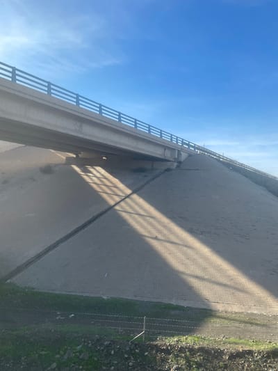 The image, correctly oriented, displays a scene looking upwards from below a large concrete bridge or overpass on a bright, sunny day. A clear, light blue sky with minimal cloud cover dominates the upper portion of the frame.
The main subject is the underside and side of the concrete bridge structure, featuring a distinctive metal railing along its edge, composed of multiple horizontal bars. The sun, positioned at an angle, casts long, prominent shadows of this railing onto the paved ground directly beneath the bridge, creating a striking pattern of parallel lines. These shadows, along with the bright light, suggest it is either morning or late afternoon.
The ground below the bridge consists of a wide, light-colored paved area, possibly made of interlocking bricks or similar material, which appears somewhat dusty or weathered. To the right of this paved area, there is a strip of natural ground with sparse green vegetation and scattered debris. A fence, appearing to be barbed wire or a similar type, runs along the boundary between the paved surface and the natural ground.
No people or vehicles are visible, and no specific activity is taking place. The concrete of the bridge shows some signs of weathering, with darker stains or discoloration visible in certain areas underneath. There is no visible text in the image. The setting is an outdoor environment, likely an infrastructure area in Jbilate, Morocco.