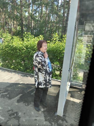 An elderly woman with auburn hair stands on a paved area next to a bus stop shelter in Zhukovsky, Russia. She is wearing a black and white floral patterned jacket, black trousers, and dark shoes. A blue scarf is visible around her neck. The bus stop shelter has a metallic frame and glass panels, displaying what appear to be schedules or advertisements. Behind the woman is a dense hedge of green bushes, and further back, a forest of tall, slender pine trees. The ground is asphalt, with some water stains and debris. The image seems to be taken from inside a vehicle, possibly through a window.