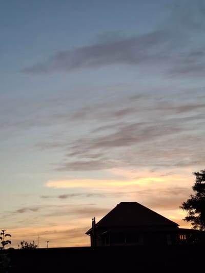 The image depicts a silhouette of a house against a twilight sky. The sky is a gradient of soft blues and oranges, with wispy clouds scattered across it. A dark-roofed house with a chimney is visible in the lower right portion of the frame, partially obscured by the silhouette of a tree. The foreground is dominated by a dark, undefined area, likely indicating a fence or the ground. The overall impression is one of a quiet evening in a residential area. There are no people or animals visible in the image.