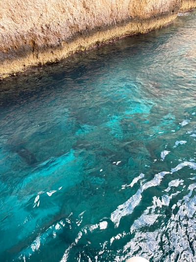 This is a high-angle, close-up shot of the clear, turquoise waters of Antipaxos, Greece, bordering a sandy, rocky coastline. The water is shallow and transparent, revealing the rippled sandy bottom beneath. Sunlight glints off the surface, creating dynamic patterns of light and shadow. The coastline on the left is textured with sand and small pebbles, with patches of green algae clinging to the rocks where they meet the water. The scene is bathed in bright daylight, suggesting a sunny day. There are no people or specific objects visible besides a hint of a white object, possibly a buoy or part of a boat, in the upper right corner. The overall impression is one of natural beauty and tranquility, characteristic of a Mediterranean beach setting.
