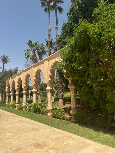 A long, low stone walkway, paved in light brown tiles, leads away from the viewer. The walkway is bordered by a row of stone arches with ornate, rounded tops, built into a wall. The arches are spaced evenly apart and are supported by pillars with decorative tops. The wall is made of smooth, light-colored stone. Behind the wall is a row of low bushes, and a grassy area. Palm trees can be seen in the background, and a dense, green bush dominates the right side of the image. The sky is clear blue. The location is in Ouled Jelal, Morocco, at coordinates 31.664, -7.872. There are no people in the scene. The image is taken from a low angle, looking toward the walkway.