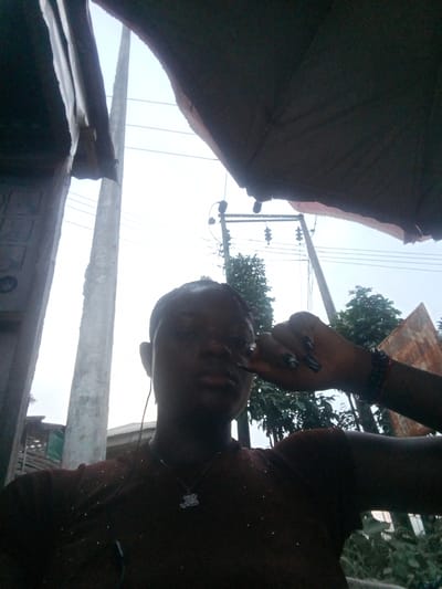 A low-angle, back-lit outdoor portrait captures a person, likely female, in Mbribit, Nigeria, during daylight hours. The subject is centered in the frame, looking towards the camera. Their face is largely in shadow against the bright, clear sky, obscuring facial features. They wear earphones, a dark-colored shirt with subtle light-colored specks, and a necklace with a small, light-colored charm, possibly a teddy bear. Their right hand, adorned with dark nail polish and a bracelet, is raised to the side of their head.
The background is dominated by a bright sky. Two large utility poles stand prominently, one on the left and another slightly right of center, supporting multiple power lines that stretch across the upper mid-ground. Lush green trees and foliage are visible beneath the power lines. Further in the background, elements of structures, including what appears to be a corrugated metal roof, can be vaguely discerned. The foreground is framed by dark, ambiguous overhead structures, possibly an awning or temporary shelter, creating a natural vignette and contributing to the strong backlighting on the subject. The scene depicts a casual moment in a public, semi-urban or residential outdoor setting. No discernible text is visible.