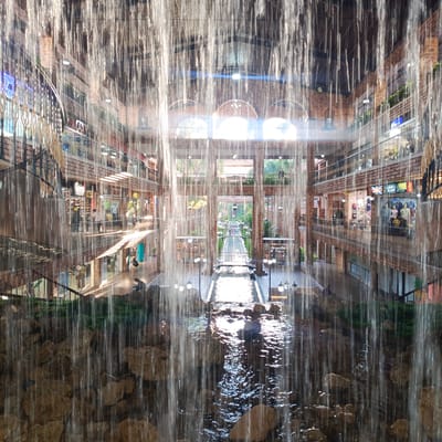 The image shows a view through a curtain of falling water into an indoor courtyard or atrium of a multi-story building. The architecture features exposed brick, arched windows, and multiple levels of walkways and shops. Sunlight streams in from the far end of the atrium, illuminating a long, narrow water channel with bridges crossing it. People are visible on the walkways, in the shops, and near the water features. The foreground is dominated by rocks and the flowing water. This location appears to be the Sambil Paraguan in Carrizal, Venezuela.