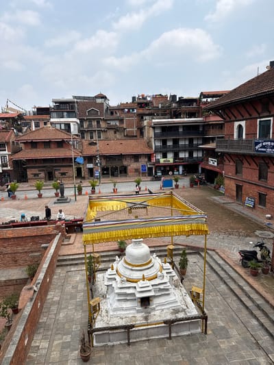 The image shows a view of Patan Durbar Square in Lalitpur, Nepal. In the foreground is a white stupa-like structure adorned with a gold-colored canopy. Several people are visible near the stupa. The background is filled with traditional brick buildings, a common architectural style in Nepal. The sky is overcast with scattered clouds.