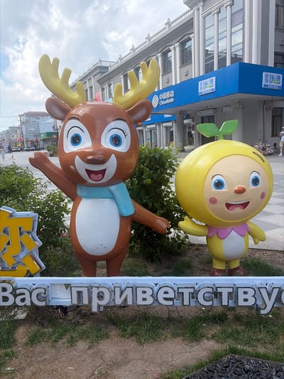 In the city of Aihui, China, a large, vibrant cartoon statue of a deer with yellow antlers stands to the left, wearing a light blue scarf. To its right is a yellow, ball-shaped cartoon character with a green sprout on its head. Both statues have wide smiles and blue eyes. Behind them, a row of buildings is visible, including a blue-fronted China Mobile store with Chinese characters. In the foreground, a white sign with large, raised silver letters spells out "Вас приветству" in Cyrillic script, suggesting a Russian influence or presence in the area. The ground is covered with dry grass and cracked earth, with some dark stones placed at the very bottom of the frame. The sky is overcast with scattered clouds.