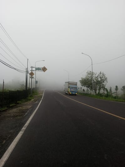 A dark asphalt road curves through heavy fog. A yellow line marks the road's center, and a white line indicates the edge. On the left, a dark fence and poles stand along the roadside. Overhead, a web of power lines stretches across the sky. A truck with a blue cab drives along the road's right side. Streetlights and warning signs are visible, obscured by the fog. Vegetation lines the roadside.