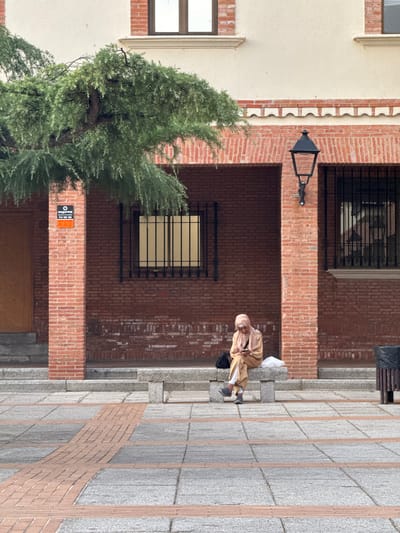 A woman wearing a light-colored headscarf and a matching outfit is sitting on a stone bench outside a brick building. She is looking down at her phone. Beside her on the bench are a black bag and a white bag. To her right, a black trash can is visible. The ground in front of the bench is paved with large gray tiles and is accented with bands of smaller orange bricks. The building behind her is made of red brick with several barred windows. A street lamp is mounted on the brick wall to the right of the woman. A large tree with green foliage hangs over the scene from the left. A security sign is affixed to a brick pillar on the left side of the image. The overall impression is an urban street scene in Las Rozas de Madrid, Spain.