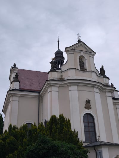 The image shows the exterior of the Church of St. Lawrence in Choceň, Czechia. The church has a Baroque architectural style, characterized by its ornate facade, curved elements, and statues of saints. A prominent feature is the tiled red roof, which contrasts with the pale yellow and white walls of the building. The church is topped with multiple spires and crosses, signifying its religious purpose. In the foreground, lush green trees partially obscure the lower portion of the church. The sky is overcast, suggesting a cloudy or diffused lighting condition. The scene is captured from a low-angle perspective, emphasizing the height and grandeur of the church.