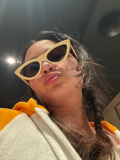 An upward-angled, close-up shot features a person, likely female, wearing sunglasses, with her head tilted sideways. The person has dark brown or black hair, which appears to be styled loosely around the face with some strands pulled back into a ponytail visible on the right side of the frame. She is wearing cream or light yellow cat-eye sunglasses with dark lenses, obscuring her eyes. Her lips are slightly parted, showing a hint of a pinkish or light red lip product.
She is dressed in a soft, fleece-like garment that is white on the outside and has orange or yellow accents, possibly a hoodie or a comfortable top. The material appears plush and cozy.
The background is predominantly dark and out of focus. In the upper left corner, a dark, circular object, possibly a ceiling speaker or recessed light fixture, is visible. Lower and slightly to the left, a bright, circular light source is present, creating a luminous point in the dark background, suggesting an indoor setting with artificial lighting. The overall lighting is dim, with the person's face illuminated by the subtle ambient light and reflections. The image has a slightly playful or stylish tone due to the pose and accessories. No text is visible in the image.