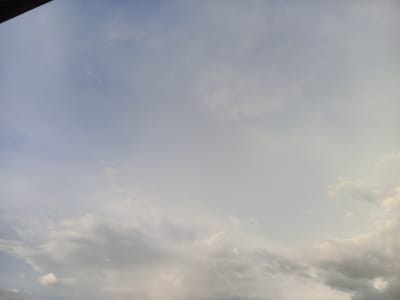 The image shows a cloudy sky with the top left corner partially obscured by a dark, horizontal object, possibly a building overhang or frame. The clouds vary in density, with some appearing wispy and translucent, while others are thicker and more opaque, casting shadows that create variations in light and texture. The lower portion of the image features more prominent, layered clouds, suggesting a transition towards overcast conditions or perhaps the setting sun's influence on the cloud formations. The lighting suggests it is either early morning or late afternoon. The scene is devoid of any human figures, animals, or man-made structures, depicting only the natural atmospheric elements. The context of Khodja Akhrar Vali, Uzbekistan, implies a wide-open sky characteristic of such a region, where such cloud formations are common.
