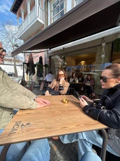 The image depicts an outdoor cafe scene on a sunny day in Chaves, Portugal. Three individuals are seated around a light-colored wooden table with a "SICAL" brand logo and a stylized female figure. A small, golden cylindrical ashtray rests on the table.
On the left, partially visible, is a male wearing glasses, a light-colored jacket, and blue jeans, with his hands clasped on the table. In the center, a female wearing brown sunglasses, a tan blazer over a patterned top, and a watch on her left wrist is looking directly towards the camera, with her right hand near her chin. A black shoulder bag is positioned next to her. On the right, a female in profile, wearing dark sunglasses and a black top, is engrossed in a smartphone held with both hands; a black bag with a braided handle is visible.
Behind the primary subjects, an outdoor seating area is covered by a brown awning. Additional dark-colored cafe tables and chairs are visible, occupied by other patrons. Through a large glass storefront, the cafe's interior reveals more customers and a warm light fixture. Hanging plants in macrame holders adorn the storefront. A sign "MÓVEL - ÓTICA" is visible under the awning.
The background shows a street with a bare deciduous tree and a parked black sedan. Historic buildings line the street, including one with a pink facade and a white balcony supported by columns, and an adjacent building featuring a light-colored stone or stucco facade with ornate windows, including a distinct quatrefoil design. The sky is clear and blue with scattered clouds, indicating bright daylight and strong shadows.