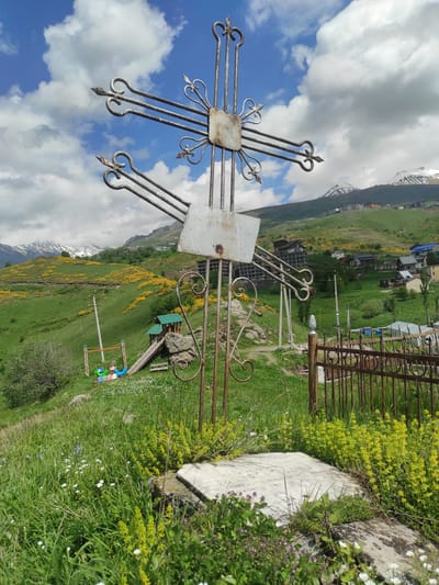 A weathered metal cross stands prominently in the foreground of an outdoor scene in Kumlistsikhe, Georgia. The cross, adorned with decorative swirls, is planted in a grassy area with wildflowers. To the left of the cross, a playground with swings and a slide is visible on a green, hilly landscape. In the background, a village with houses and apartment buildings is nestled on the slopes of mountains, some of which are capped with snow. The sky above is a mixture of blue with scattered white clouds. The overall scene depicts a serene, mountainous rural setting with elements of both religious and recreational life.