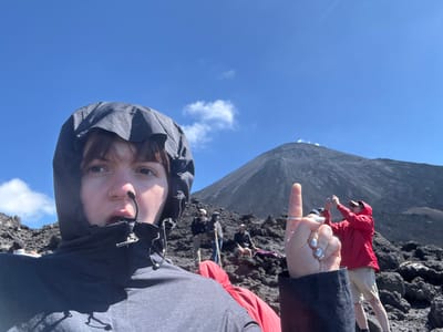 A selfie is taken looking downward with a blue sky and some scattered clouds in the background. The photographer wears a hooded gray jacket. In the distance, a large, rocky mountain dominates the mid-ground. To the left, a person in a red jacket is partially visible. Another person's legs are visible in the upper left corner. The photographer gestures with a raised index finger. Other indistinct figures can be seen on the rocky ground.
