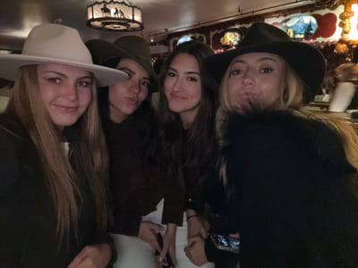 Four young women are closely posed, their faces close together. They are inside a building. Each one is wearing a hat. The woman on the left wears a dark hat. The woman on the right wears a white hat. In the center, the woman wear a dark hat. Behind them is an ornate ceiling.