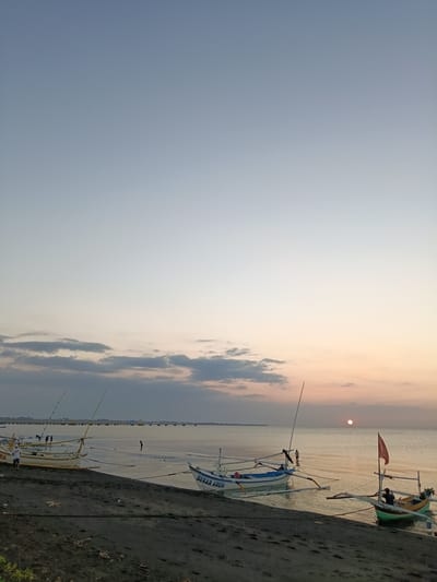 The image depicts a serene coastal scene at dusk in Jangkar, Indonesia. The foreground is a dark, sandy beach with several traditional wooden fishing boats moored near the shore. The boats, of varying sizes and colors, are characterized by their outriggers and tall masts. One boat in the middle ground has the visible text "SEKAR ARUM" painted on its side.
In the water, a few people are present. One person stands in the shallow water near a boat, while others are further out, possibly fishing or preparing their vessels. In the distance, a long pier or breakwater stretches across the horizon, with structures visible on it.
The sky transitions from a pale blue at the top to softer hues of pink and orange near the horizon, indicating either sunrise or sunset. A small, reddish sun is visible low on the horizon, casting a warm glow on the water. The sea is calm, with gentle ripples reflecting the sky's colors. The overall mood is peaceful and tranquil, capturing the quiet beauty of a coastal evening. The weather appears calm, with a few scattered clouds adding texture to the sky.
