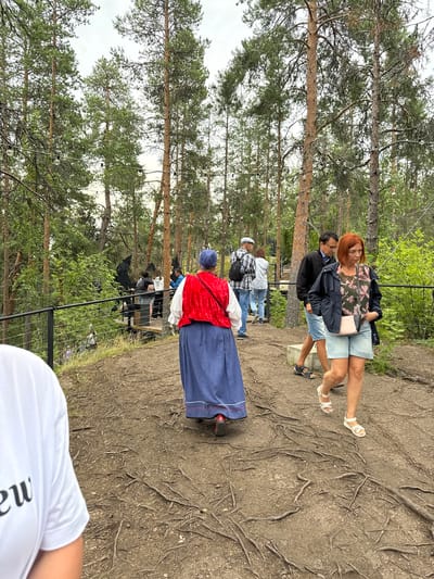 In Kosalma, Russia, a group of people are walking on a dirt path winding through a forest with tall pine trees. Some of the trees have lights strung between them. In the foreground, a person wearing a white t-shirt with black lettering is partially visible. To the right, a woman with short red hair, wearing a floral-patterned top and denim shorts, walks forward. Behind her, a man in a dark jacket and denim shorts is walking. Further back, a woman in traditional folk attire, a red vest over a white shirt and a blue skirt, is walking away from the camera. Other people are seen on a wooden boardwalk in the background. The ground is covered with exposed tree roots.