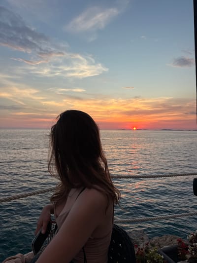 The image captures a young woman with long, dark brown hair, viewed from behind, as she gazes out at a sunset over the Adriatic Sea in Primošten, Croatia. She is wearing a light-colored, ruched dress and is seated, with her left arm resting on a railing. The sea is a deep blue, with gentle waves reflecting the warm hues of the setting sun. In the distance, a sliver of land or islands is visible on the horizon. The sky is a gradient of colors, transitioning from a pale blue at the top to shades of orange, pink, and yellow near the horizon. A rope railing runs horizontally across the foreground, separating the seating area from the sea. The scene evokes a sense of tranquility and beauty, characteristic of a Mediterranean sunset.