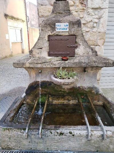 An old stone public drinking fountain, labeled "Eau Potable" (Drinking Water) in French, features a metal door above a decorative plant in a pot. Water flows from a metal faucet into a mossy stone basin divided into sections by metal bars. The fountain is situated on a narrow stone street, adjacent to buildings with white siding and a closed shutter.
