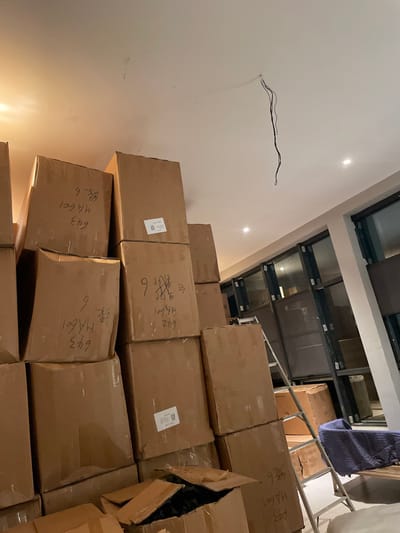 The image shows a stack of cardboard boxes reaching towards the ceiling. Many boxes are labeled with handwritten characters, including "643 HA601 绿6". A few boxes are partially open. Electrical wiring extends from the wall. Above the boxes, a series of glass panels are visible in the ceiling, along with a ladder and a draped object. The overall scene suggests a storage area or delivery location.