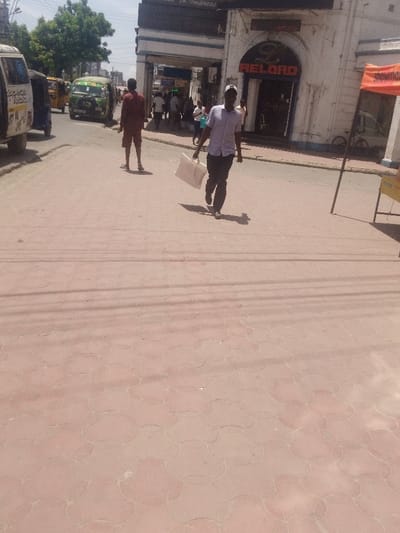 The scene depicts a street view in Majengo ward, Kenya, on a clear, sunny day. The foreground consists of reddish-brown interlocking paving stones with a repeating circular pattern, exhibiting visible shadows cast from the left, indicating bright overhead sunlight.

Two adult males are prominent in the midground. One individual, on the right, walks forward toward the viewer, carrying a large, light-colored rectangular bag with two handles in his left hand. He wears a light-colored collared shirt, dark trousers, and a white baseball cap. His shadow extends behind him. A second individual, on the left, stands barefoot in the street, facing away from the camera, wearing a dark reddish-brown shirt and shorts.

On the left side of the street, several vehicles are present: a partially visible white van with visible markings on its rear, a yellow tuk-tuk behind the van, and a green matatu (minibus) with a dark front bumper further down the road.

On the right side of the street, a multi-story building with a white facade and blue trim is visible. It features an arched entrance with the word "RELOAD" displayed in red, illuminated letters above the doorway. Adjacent to this, another section of a building has an archway with "Empress" partially legible above it. Multiple blurred individuals are visible near these building entrances. An orange banner or tent structure on poles is positioned on the far right, partially obscuring a white bicycle and a yellow sign. Green foliage is visible in the distant background on the left.