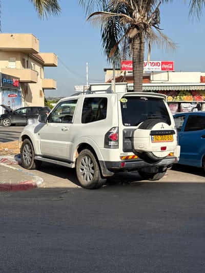 A white Mitsubishi Pajero SUV is parked on a paved road in Jaljulia, Israel. The car is facing the camera and is parked on the right side of the road. The car has a license plate that reads "92-360-63." There is a palm tree to the right of the car. The car is in front of a building with a red Coca-Cola sign. There is another car partially visible in the background on the left, as well as a building with a red roof and a red sign. The road is mostly empty, with no people visible. The weather is clear and sunny.