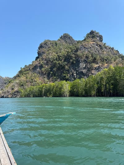 A rocky island with trees stands beside calm turquoise water under a bright blue sky. The island's terrain is rugged, with patches of greenery clinging to its surface. The water's surface reflects sunlight, creating a shimmering effect. A portion of a boat is visible in the foreground, with a wooden deck and a blue-painted section. The scene evokes a sense of tranquility and natural beauty.