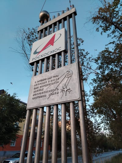 This is a low-angle, outdoor, medium shot of a monument dedicated to the launch of Sputnik 1. The monument features a spherical object, representing Sputnik, at its apex, supported by vertical metal poles. Below this, a sign displays the "Roscosmos" logo, which includes a stylized rocket and a red arrow. Further down, a plaque is inscribed with text in Russian, commemorating the first launch of an artificial satellite of Earth on October 4, 1957. The monument is set against a clear blue sky, with trees framing the scene. In the background, residential buildings and a road are visible, suggesting an urban or semi-urban setting. The lighting indicates it is either early morning or late afternoon. The overall mood is one of historical commemoration and technological achievement. The text on the plaque reads: "4 ОКТЯБРЯ 1957 ГОДА ВПЕРВЫЕ В МИРЕ БЫЛ ОСУЩЕСТВЛЕН ЗАПУСК ИСКУССТВЕННОГО СПУТНИКА ЗЕМЛИ" which translates to "ON OCTOBER 4, 1957, FOR THE FIRST TIME IN THE WORLD, THE LAUNCH OF AN ARTIFICIAL EARTH SATELLITE WAS CARRIED OUT."