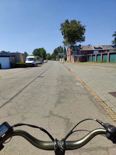 The image depicts a street scene in Breda, Netherlands, taken from the perspective of someone riding a bicycle. The street is paved with asphalt and shows some signs of wear. To the left of the street, there are buildings with garages and a white van parked. To the right, there is a sidewalk paved with light-colored rectangular tiles, bordered by a row of red bricks. Further down the street, there are houses with tiled roofs and trees lining the sidewalk. The sky is clear and blue, indicating a sunny day. The foreground of the image shows the handlebars of the bicycle, with a digital display on the left. The overall environment suggests a residential or mixed-use area within the city.