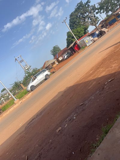 The image depicts a roadside scene in Kassa, Nigeria. A silver minivan is parked on the right side of a dirt road. To the left of the road, a power pole with transformers and wires stands tall. Several small buildings line the opposite side of the road, some with corrugated metal roofs. A large tree with lush green foliage is visible behind the buildings. The sky is a clear blue with scattered white clouds. The ground is a reddish-brown dirt, with some debris and vegetation visible along the roadside. The scene appears to be a typical day in a Nigerian town, with signs of daily life and infrastructure.