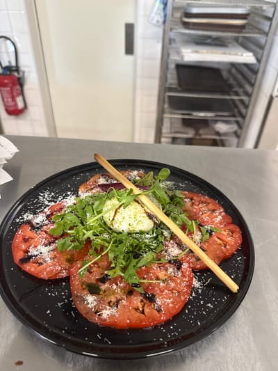 This image shows a close-up, overhead view of a Caprese salad served on a black plate. The salad features large, sliced tomatoes, fresh arugula, a ball of mozzarella cheese, and a breadstick. The dish is sprinkled with grated cheese and drizzled with a dark sauce, possibly balsamic glaze. The salad is presented on a stainless steel countertop. In the background, to the left, a commercial kitchen rack holds various metal trays and containers, suggesting a restaurant or food service setting. Further in the background, a white tiled wall is visible, with a red fire extinguisher mounted on it. The lighting is bright, indicating an indoor setting, likely during the day. The overall impression is a professionally prepared and appetizing dish. No people are visible in the image. The location context provided is Rivesaltes, France.
