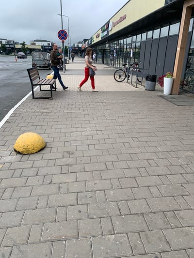 In Novoe Shigaleevo, Russia, a father carries a baby while walking on a cobblestone sidewalk in front of a row of shops. A woman in red pants walks past them in the opposite direction. A bicycle is parked at a bike rack near the shops, and a trash can and a potted plant are also visible. A yellow concrete barrier is on the sidewalk. The sky is overcast.