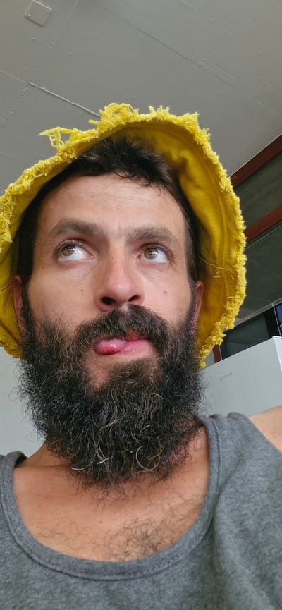 A man with a beard and a yellow hat is sticking his tongue out while looking up and to the side. He is wearing a grey tank top. The man is indoors, with a white ceiling above him. There is a refrigerator behind him, with the brand name "COLDTEC" visible. The image was taken in Uetendorf, Switzerland.