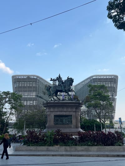 The image captures a bronze equestrian statue in front of a modern, curved building, likely located in San Salvador, El Salvador. The statue sits atop a stone pedestal adorned with relief carvings. In the foreground, there are plants with red and green leaves, and a paved walkway is visible. In the background, a building with a distinctive curved design and a grid-like facade is apparent. The sky is blue with some clouds. A utility wire runs across the upper left portion of the image. People are seated at the base of the statue, and a person wearing all black is walking.