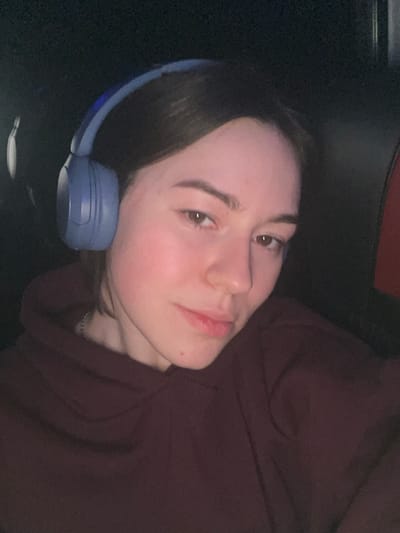 An indoor, medium close-up shot features a young person, likely female, with fair skin and dark hair, wearing blue over-ear headphones. Her head is tilted, suggesting she might be lying down or reclining, looking directly at the camera with a neutral to slightly contemplative expression. Her cheeks show a natural flush. She is wearing a dark, possibly maroon or dark red, hoodie or top. The background is very dark and out of focus, providing minimal details about the setting beyond suggesting an indoor environment with low lighting, possibly evening or night. There are no discernible location cues specific to Palanca, Moldova, within the image itself. No text is visible.